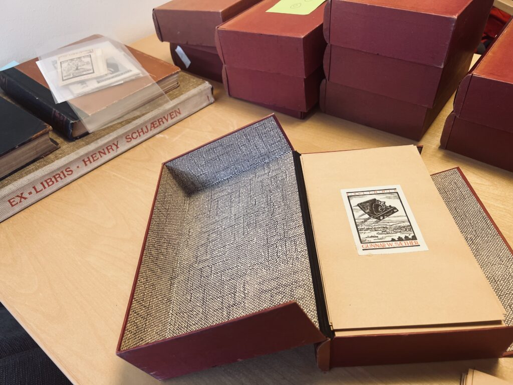 A snapshot of boxes with exlibris in them. They lay on a table. One box is open and an exlibris is visible. Books to the left.